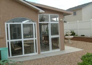 A tan house featuring a small glass-enclosed porch with an arched window and a white-framed glass door leading to a gravel patio